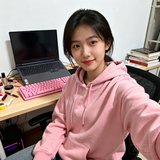 一位20岁出头的女性玩家在宿舍书桌前自拍，穿着粉色连帽卫衣，身后可以看到笔记本电脑和粉色键盘